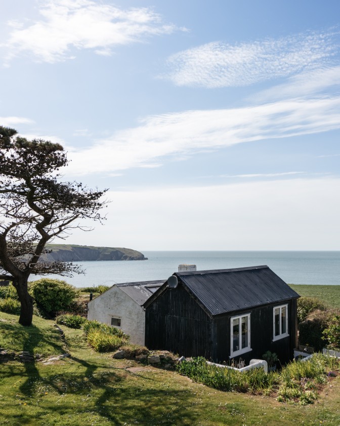 A black wooden bothy on grass with views over cliffs and ocean