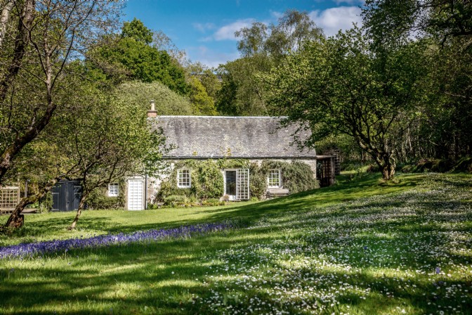 In a flowered lawn surrounded by trees is white, ivy-covered cottage Little Eden on Loch Lomond, Scotland