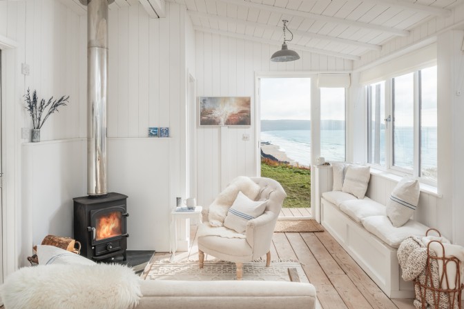 GP2596 - A white-washed living room with sofas and woodburner, and an open door to the cliffs overlooking sea