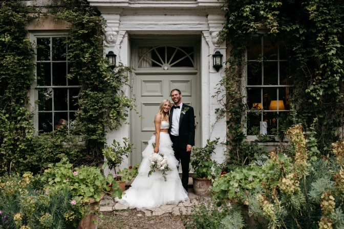A bride and groom stand in front of a doorway surrounded by greenery