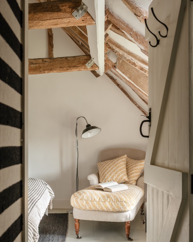 A white bedroom with crooked timber beams, chair with footstall and floor lamp