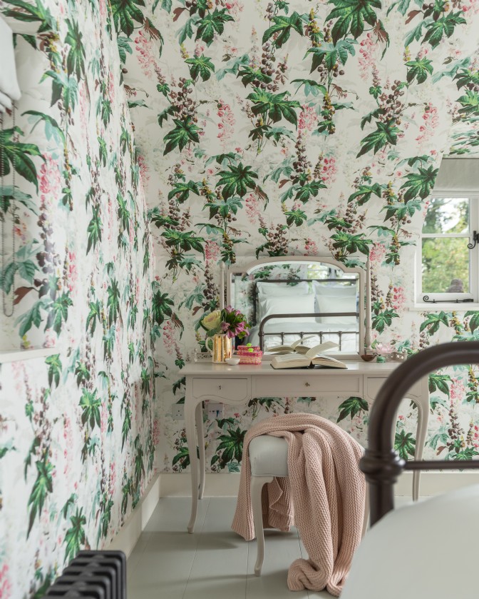 A bedroom covered in blousy wallpaper by House of Hackney, a white vanity dresser and stool with blanket thrown over