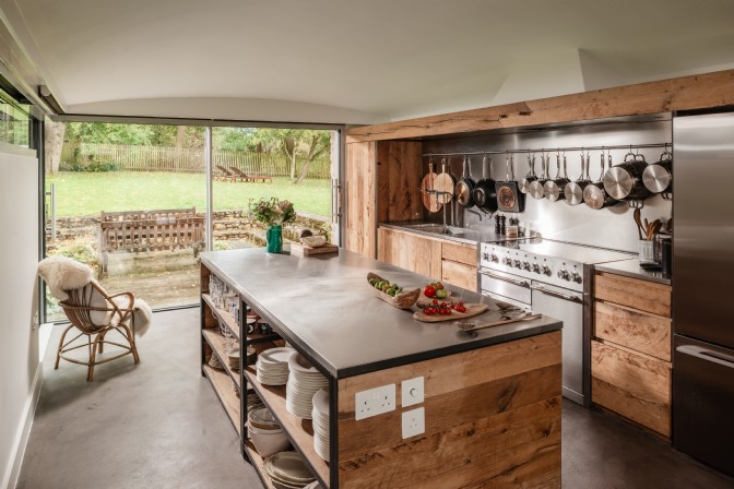 A rustic kitchen with island, wooden cabinets and large windows looking out across the garden
