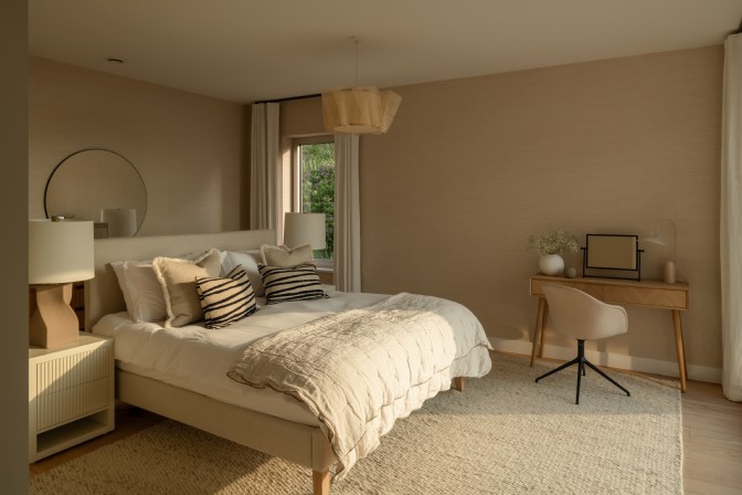 An image of a neutral bedroom with large bed, desk, bedside tables and mirror