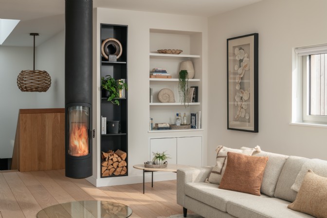 An image of a living space with lit woodburner, sofa, wood store and shelves filled with coastal trinkets