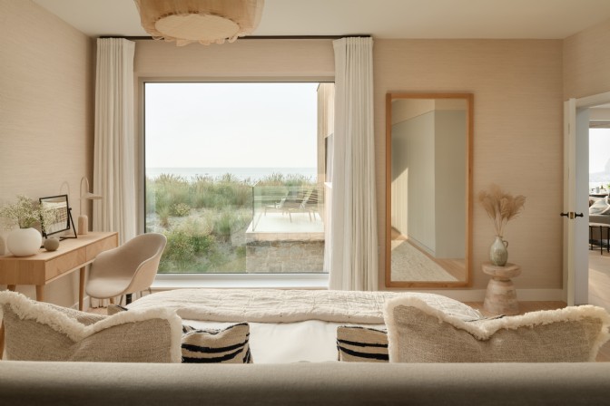An image from a bed looking out across the beach and sea. The windows are large and the colour scheme in the room is neutral. There is a desk with chair and mirror.