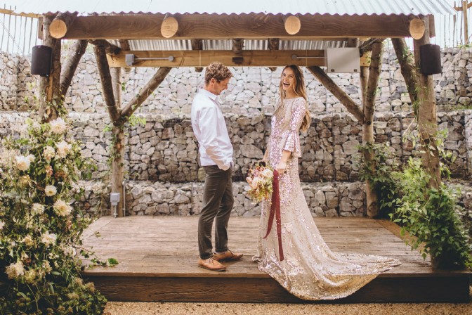 An image of a couple getting married at their relaxed wedding. There are flowers climbing up the pergola and the bride is laughing.