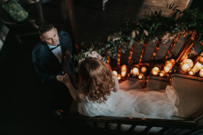 An image of a wedding couple walking down some stairs. There are candles lit and foliage wrapped around the staircase. The bride wears a white dress and the groom a suit with a bowtie. 
