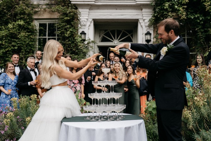 An image of a bride and groom filling a Champagne tower. The bride's dress is two piece and there is a crowd of smiling wedding guests behind them