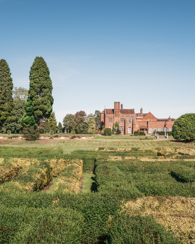 A red brick manor houses in the distance, seen from a topiary lawn maze