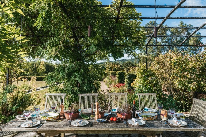 An image a table under a pergola covered in greenery. The table is set up with plates, glasses and plants in terracotta pots.