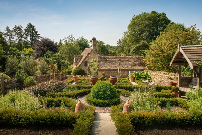 An image of a cottage garden; there are terracotta rhubarb forcers, bamboo poles, and a Cotswold stone cottage.