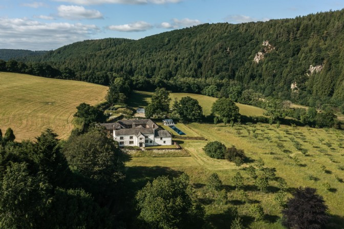 An image of a remote white farmhouse in a valley surrounded by trees, with an orchard spreading out in front of it